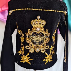 May include: A black denim jacket with a gold and rhinestone embellished design. The design features a crown, a shield, and floral accents.