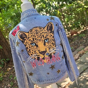 Könnte beinhalten: Eine hellblaue Jeansjacke mit einer großen Leopardenkopf-Grafik auf der Rückseite mit blauen Augen. Das Design enthält den Text "WILD THANG" und "ROAR" in Rot, akzentuiert mit silbernen Strasssteinen, sternförmigen Aufnähern und langen silbernen Fransen.