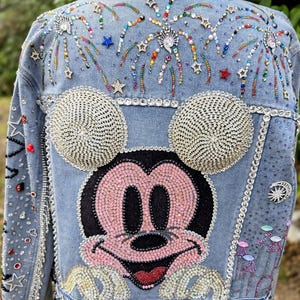 Könnte beinhalten: Hellblaue Jeansjacke mit großem Mickey Mouse-Gesicht aus rosa und schwarzen Strasssteinen. Die Jacke ist mit funkelnden Perlen, feuerwerksartigen Designs und einem dekorativen Besatz verziert.