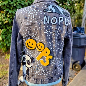 May include: A dark gray denim jacket embellished with rhinestones and patches. The patches include a panda, two smiley faces, and the word "OOPS" in yellow. The word "NOPE" is also featured on the back of the jacket.