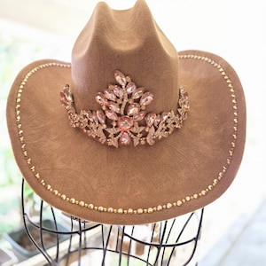 May include: A tan cowboy hat featuring a subtle paisley pattern embossed on the felt material. The front is adorned with a large, ornate tiara-style embellishment made of light pink crystals. The brim is lined with a row of small gold beads.