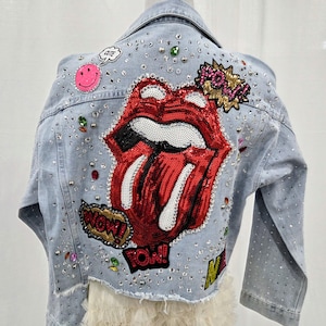 Può includere: Giacca di jeans azzurra decorata con strass e toppe. Presenta un grande design di labbra rosse con paillettes, una faccina sorridente rosa e le parole "WOW!" e "CUTE" in fumetti. Un capo di moda.