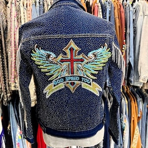 May include: A dark blue denim jacket, heavily embellished with rhinestones. The back is adorned with a colourful embroidered design of a cross, wings, and the text "GOD SPEED YOU". The sleeves have fringe detailing.