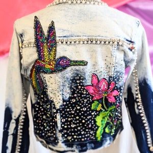 Può includere: Una giacca di jeans blu chiaro con ricamo di colibrì e fiore colorato. La giacca è decorata con strass e perle.