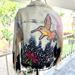 Peut inclure: Une veste en jean délavé clair ornée au dos d'un grand motif de colibri et de fleurs en strass colorés. La veste est agrémentée de cristaux irisés dispersés et de longues franges métalliques dorées fixées sur les manches.