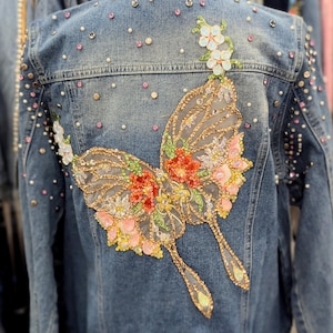 May include: A denim jacket with a large, embellished butterfly design on the back. The butterfly features colorful floral accents and sparkling rhinestones. The jacket is adorned with additional rhinestones and pearls.