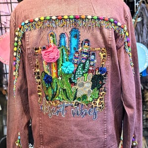 May include: Dusty rose denim jacket with colorful beaded trim and a desert-themed design on the back. The design features cacti, flowers, and the text "desert vibes". The jacket has a button closure and decorative embellishments.