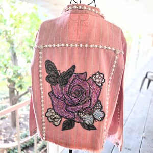 Puede incluir: Una chaqueta vaquera de color rosa empolvado con rayas verticales de pedrería, cuello ribeteado con perlas blancas y una gran aplicación en la espalda de una rosa morada rodeada de mariposas negras e iridiscentes, decorada con cuentas y cristales.