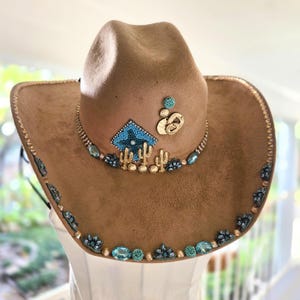 May include: A tan cowboy hat featuring a decorative band adorned with turquoise and gold-toned embellishments, including small cactus figures, a diamond-shaped beaded patch, and various blue and metallic accents along the brim and crown.