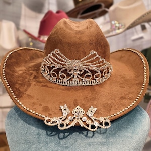 May include: A brown suede cowboy hat with a rhinestone trim and a decorative rhinestone tiara. The hat has a wide brim and a classic cowboy hat shape. The tiara is centered on the front of the hat.