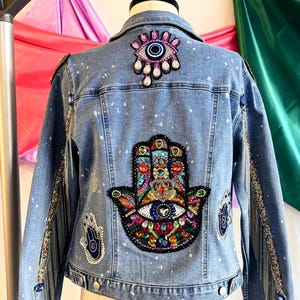 May include: A blue denim jacket with a hand-shaped patch with colorful jewels and an evil eye design. The jacket has a white splatter paint design and beaded fringe on the sleeves.