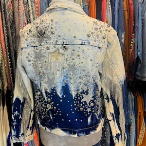 May include: Light wash denim jacket with a unique design. The jacket features a dark blue and white tie-dye effect, embellished with silver studs, rhinestones, and star-shaped accents. Silver fringe hangs from the sleeves, adding a touch of glamour.