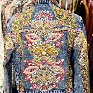 May include: A denim jacket with intricate embroidery and embellishments. The jacket features a floral design with pink, yellow, and blue crystals and beads.