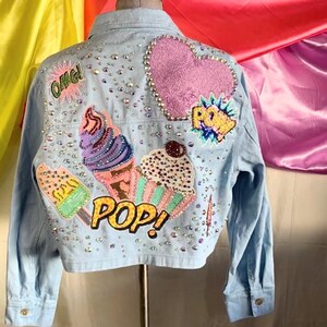Op de afbeelding: Een lichtblauw spijkerjack met een achterpand versierd met verspreide strass-steentjes, een grote roze hartvormige patch en geborduurde patches van een ijshoorntje, een cupcake, een ijsje en de tekst "OMG", "POW", "POP".