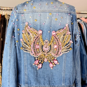 May include: A light-wash denim jacket with a large, ornate beaded design on the back. The design features pink and gold embellishments in a stylized wing or bird shape. The jacket is also adorned with scattered pink, gold, and silver studs.