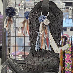 Op de afbeelding: Een zwarte leren cowboylaars met ingewikkelde uitgesneden patronen en paillettenversieringen. Aan de treklus hangt een decoratieve bedel bestaande uit een blauwe stoffen roos, een kristallen ster, diverse veren en een wit lint met de tekst CHANEL.