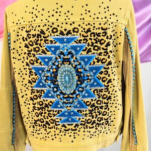 May include: A yellow denim jacket with a blue and black geometric design on the back. The design features a leopard print and rhinestones. The jacket has fringe on the sleeves.