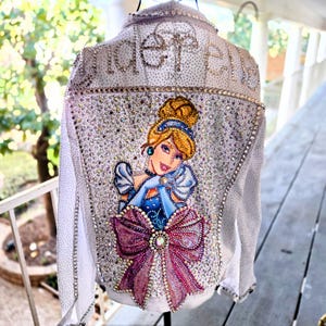 May include: A white denim jacket embellished with sparkling rhinestones and a Cinderella illustration on the back. The jacket features a large, jeweled bow and the word "Cinderella" spelled out in rhinestones across the top.