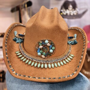 May include: A brown cowboy hat with elaborate embellishments. The hat features a decorative band with turquoise and clear gemstones, a central brooch with blue and green stones, and turquoise tassels. The brim is also adorned with gemstones.