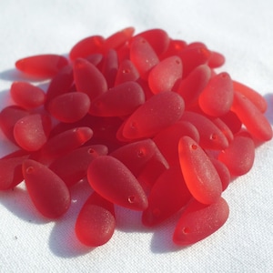 May include: A pile of red, teardrop-shaped beads with a matte finish. Each bead has a small hole at the top for stringing. The beads are a vibrant, solid red color and appear to be made of glass or a similar material. They are scattered on a white surface.