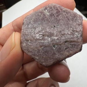 May include: A rough, hexagonal, purple-grey gemstone with a striated surface.