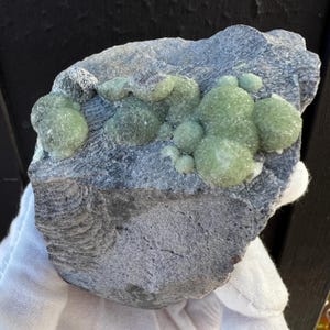Puede incluir: Un grupo de cristales de prehnita verdes sobre una matriz gris oscura. Los cristales están agrupados en forma redondeada y tienen una superficie lisa y vidriosa.