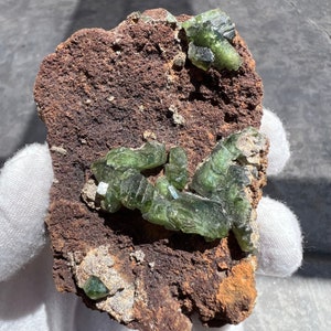 May include: A cluster of green, translucent crystals on a brown, rough rock. The crystals are sharp and have a glassy appearance.