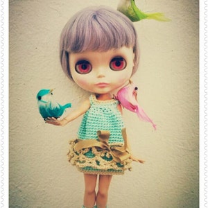 May include: A doll with purple hair and red eyes wearing a crocheted dress with a brown ribbon and blue and pink bird accessories.