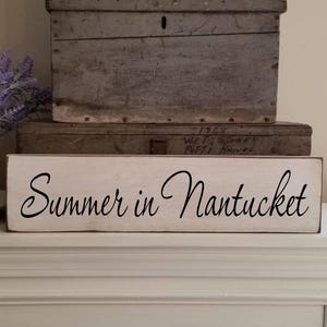May include: A white wooden sign with the words "Summer in Nantucket" in black script. The sign is on a white mantel, with a white pitcher of purple flowers and two weathered wooden boxes in the background.