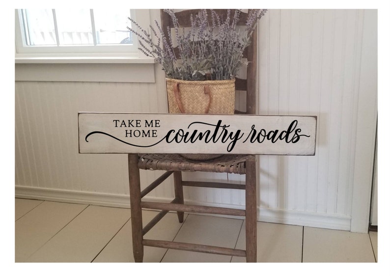 May include: White wooden sign with black text that reads "Take Me Home country roads". The sign is sitting on a wooden chair with a wicker seat.