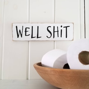 May include: A wooden sign with the words "WELL SHIT" in black lettering hangs above a wooden bowl filled with white toilet paper rolls. The sign is rectangular with a white background and a distressed edge.