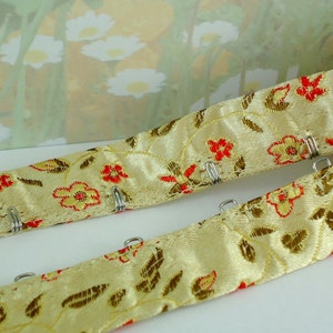 May include: A pair of beige satin ribbon with red floral print and silver metal hooks. The ribbon is used for sewing and crafting.