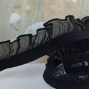 May include: Black elastic trim with a ruffled mesh overlay. The trim is folded in half, showing the elastic backing.