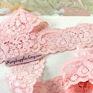 May include: Close-up of delicate, light pink lace ribbon. The lace features an intricate floral pattern with scalloped edges. The ribbon is folded and layered, showcasing its texture. The text "MarySupplies.Etsy.com" is visible on a small tag.