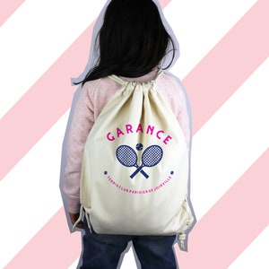 Può includere: Uno zaino bianco con coulisse con un design di racchetta da tennis e palla rosa e blu. Il testo "GARANCE TENNIS CLUB PARISIEN DE JOINVILLE" è stampato sullo zaino.
