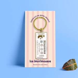 Può includere: Portachiavi in metallo dorato a forma di biglietto della metropolitana parigina. Il biglietto riporta la scritta "METROPOLITAIN" e "GOLD TICKET". Il portachiavi è attaccato a un anello in metallo dorato. La carta è su uno sfondo a strisce rosa e bianche con il testo "los imprimousos" e "Dessiné à la main à Paris".