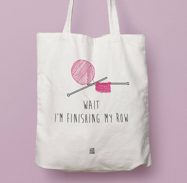 K&ouml;nnte beinhalten: Wei&szlig;e Einkaufstasche mit einer rosa Wollkugel und Stricknadeln. Der Text auf der Tasche lautet "Wait I'm finishing my row". Am unteren Rand der Tasche befindet sich ein kleines Logo mit der Aufschrift "The Other Fish".