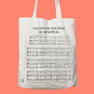 May include: A white tote bag with black text that reads "LEÇONS DE SOLFÈGE DE JOSEPH B." and features a sheet of music with musical notes. The bag has a black logo that reads "THE OTHER FISH".