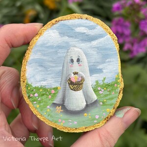 May include: A hand-painted wooden slice depicting a white ghost holding a basket of colourful eggs. The ghost has a sweet, cartoon-like face and is standing in a field of green grass with pink and yellow flowers.