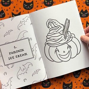 Pu&ograve; includere: Illustrazione in bianco e nero di una zucca con gelato, da un libro da colorare a tema Halloween. Il testo "1. PUMPKIN ICE CREAM" &egrave; in un riquadro sulla pagina sinistra. Lo sfondo &egrave; arancione con motivi di gatti neri e stelle.