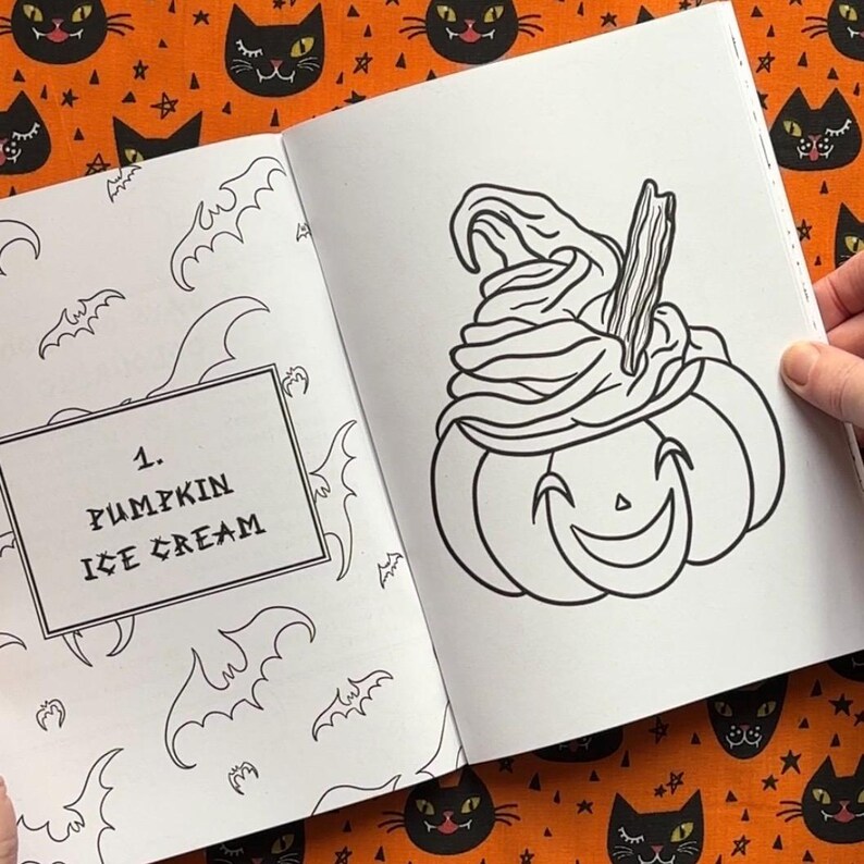 Pu&ograve; includere: Illustrazione in bianco e nero di una zucca con gelato, da un libro da colorare a tema Halloween. Il testo "1. PUMPKIN ICE CREAM" &egrave; in un riquadro sulla pagina sinistra. Lo sfondo &egrave; arancione con motivi di gatti neri e stelle.