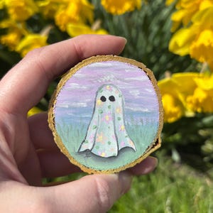 May include: A hand-painted wooden round ornament featuring a ghost with a floral pattern against a purple and blue sky background.