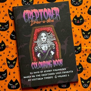 Pu&ograve; includere: Un "Creptober Draw-a-thon Colouring Book" a tema spettrale. La copertina presenta una donna in una bara, con il testo "31 Days of Spooky Colouring" e "Volume 1". Il libro &egrave; ambientato su un tessuto arancione con motivi di gatti neri e stelle.