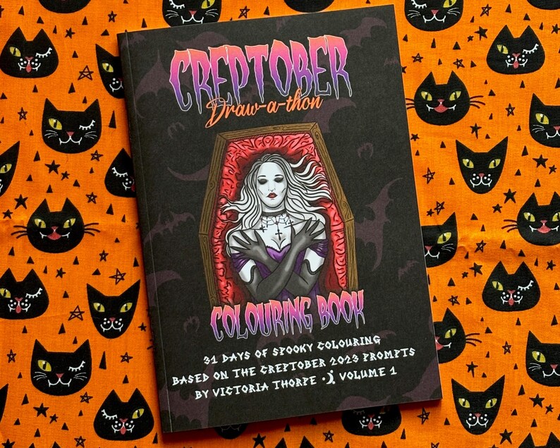 Pu&ograve; includere: Un "Creptober Draw-a-thon Colouring Book" a tema spettrale. La copertina presenta una donna in una bara, con il testo "31 Days of Spooky Colouring" e "Volume 1". Il libro &egrave; ambientato su un tessuto arancione con motivi di gatti neri e stelle.