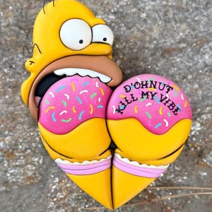 May include: A novelty item featuring a yellow cartoon character with a surprised expression, biting into a heart-shaped donut. The donut is pink with colorful sprinkles and the text "D'ohnut Kill My Vibe".