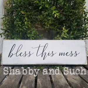 May include: White wooden sign with black calligraphy that reads "bless this mess". The sign is distressed and has a rustic look. The sign is sitting on a wooden surface.