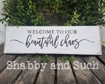 May include: White wooden sign with black text that reads "Welcome to our beautiful chaos". The sign has a distressed, rustic look and is sitting on a wooden surface.