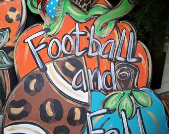 May include: Wooden pumpkin-shaped door hanger with the words "Football and Fall" in white. The pumpkin is painted orange, brown, and teal, with a decorative bow featuring football and leopard print ribbons. A festive fall decoration.