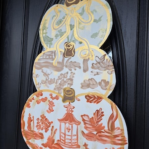 May include: A decorative wooden pumpkin-shaped door hanging. The three stacked pumpkin shapes are painted with a white background and gold trim. Each pumpkin has a different design in shades of brown and orange, with a gold bow at the top.