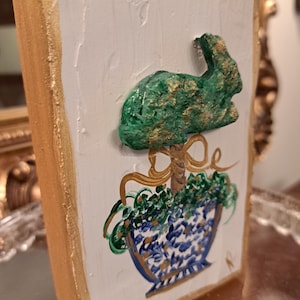 May include: A rectangular, hand-painted wooden plaque with a gold-painted frame. The white surface features a green bunny and a blue and white floral pot design. Gold accents are used throughout the artwork.